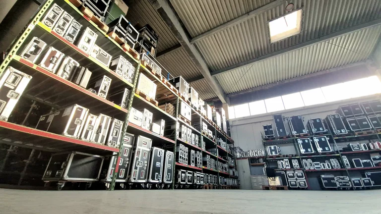 Ein Blick in eine Lagerhalle, aus der Bodenperspektive schräg hoch fotografiert. Alle Regale sind mit 3 bis 4 Ebenen übereinander voll mit verschiedenen Cases. Durch ein Oberlicht im Dach und eine Fensterreihe oberhalb der Wand strahlt das Sonnenlicht herein.