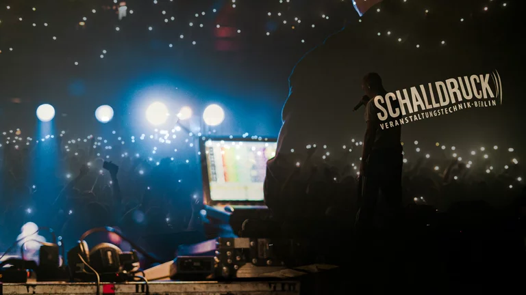 Ein Kombi-Bild, überblended aus einer Person an der Technik vorn rechts, mit schwarzem Pullover und Schalldruck-Logo auf dem Rücken, und Publikum welches Feuerzeuge hält. Es sieht aus wie ein Sternenhimmel.