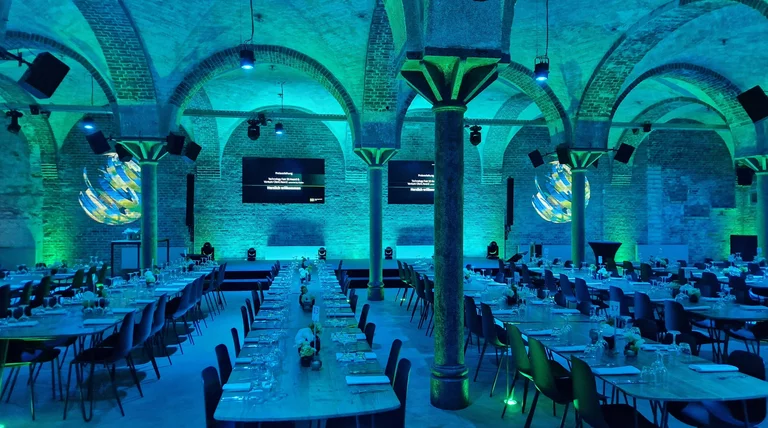 Der Gewölbekeller der Eventlocation Deep, mit langen Dinnertischen, alles in blaugrünes Licht getaucht. In der Mitte sieht man eine alte runde Säule, welche die Gewölbedecke mit den vielen Rundbögen trägt. Hinten an der Wand sieht man 2 große flache Bildschirme an der Wand, links und rechts davon an die Wand projezierte runde verwischte Grafiken. Die Dinnertische sind fein gedeckt.
