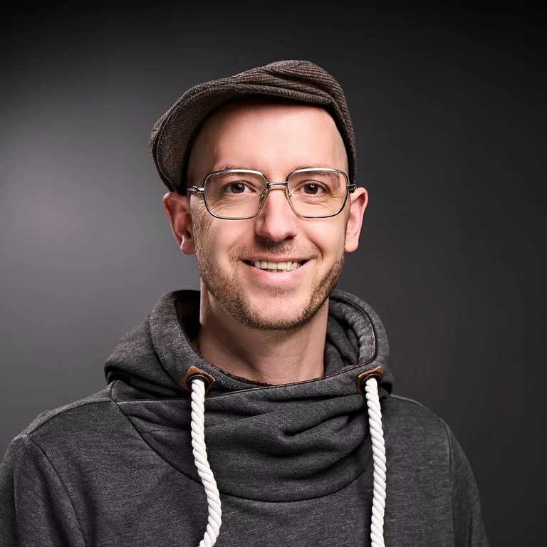 Portrait-Foto von Ingo König, Projektleigung, in einem dunkelgrauen Pullover und mit Schiebermütze auf dem Kopf. Er trägt eine Brille. Er lächelt.