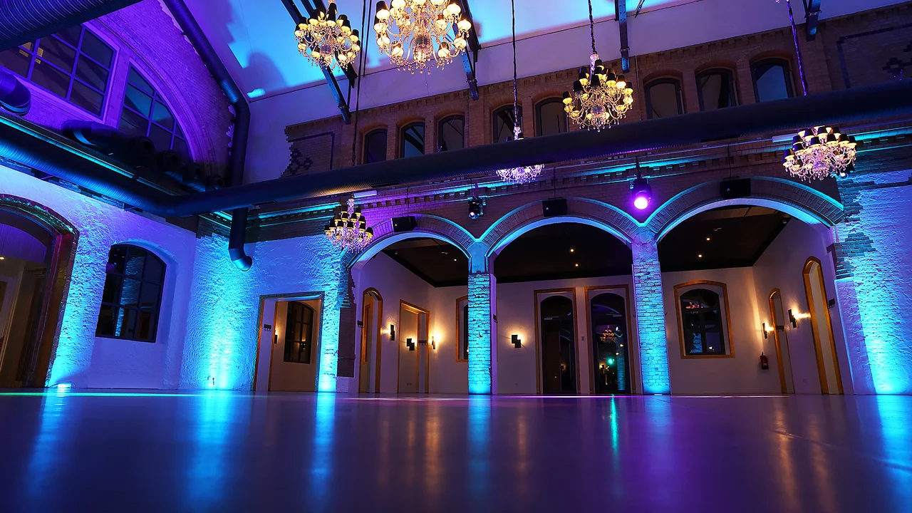 Dicht vom Boden aus schräg hochfotografiert, man sieht die 3 Rundbögen und die 2 Mittelsäulen der Wartehalle, ein altes Bahnhofsgemäuer. Alles ist hellblau beleuchtet, der glatte Boden spiegelt die Lichter wider. Von oben kommen die hängenden Kronleuchter in gelblich-weissem Licht.