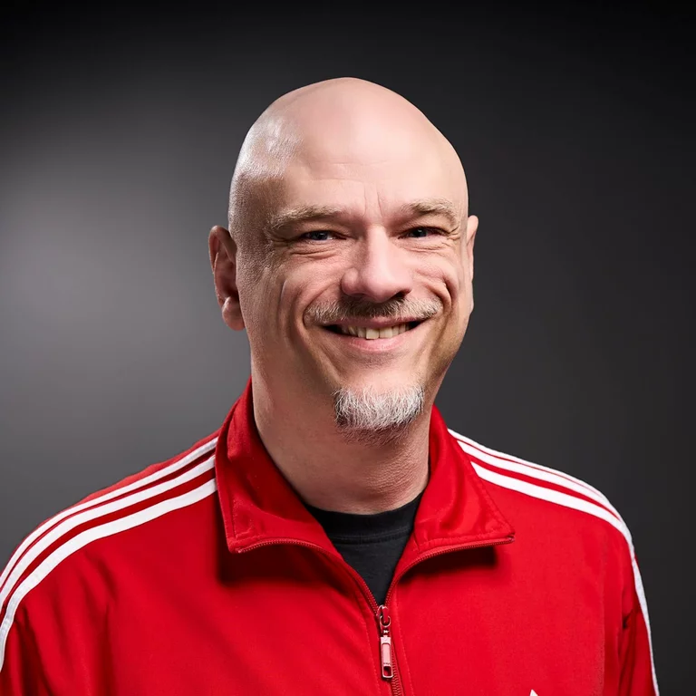 Portrait-Foto von Thomas Tieke, Projektleitung & Disposition, in einer roten Adidas-Jacke mit weissen Streifen. Er hat einen Kinnbart und lacht.